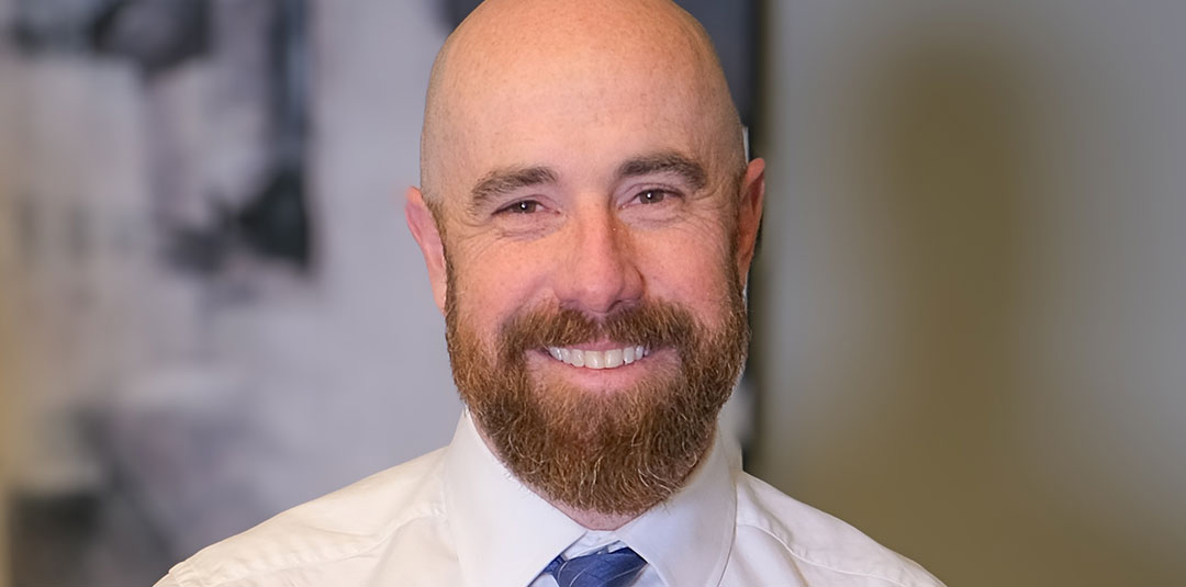 Professional man standing behind a desk wearing a dress shirt and tie in a modern office setting.