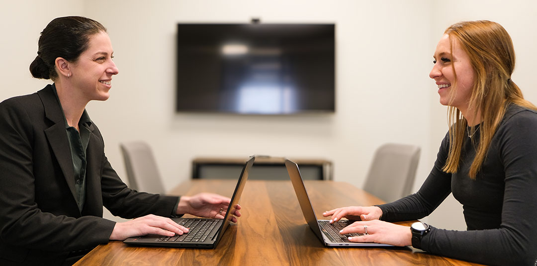 Two people meeting across a table while working on laptops in an office.