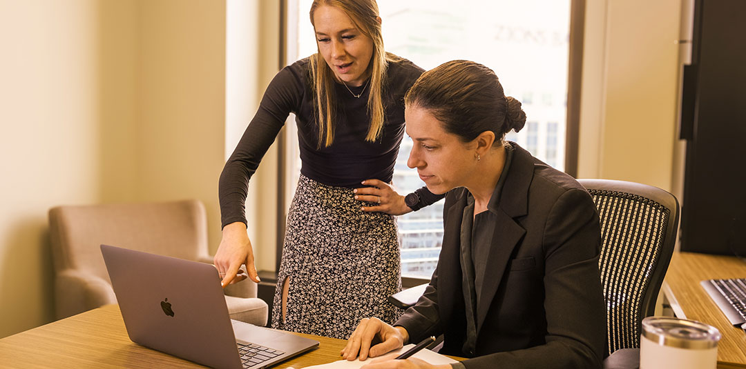 Two people reviewing information on a laptop while taking notes at a desk.
